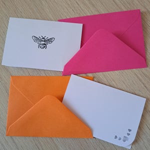 May include: Two white rectangular note cards and two envelopes in bright pink and orange. One card features a black ink illustration of a bee, while the other card has a rounded corner with small heart-shaped cutouts in the bottom right corner.