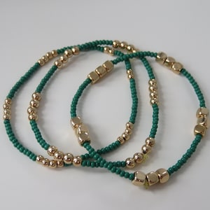 May include: Three green and gold beaded bracelets with gold square beads. The bracelets are stacked on top of each other.