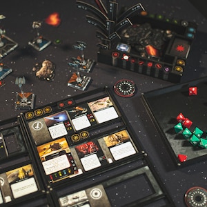 Może przedstawiać: Gra planszowa w tematyce Gwiezdnych Wojen z czarną i szarą planszą, czerwonymi i zielonymi kostkami oraz miniaturowymi statkami kosmicznymi. Plansza do gry ma wiele sekcji z kartami i żetonami.