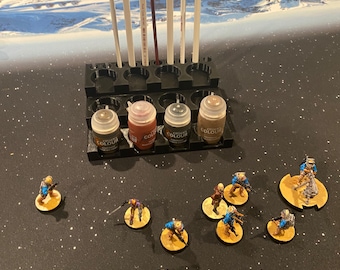May include: A black paint rack with nine paint bottles, four of which are brown, and five paintbrushes.  The rack is on a black table with a white and blue background.  There are eight miniature figures on the table, all wearing blue and brown clothing.