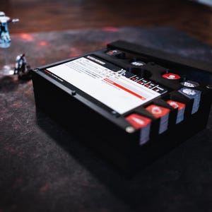 May include: A black dice box with a clear lid and red and white dice. The box has a compartment for storing dice and a separate compartment for storing cards. The box is open and the dice are visible.