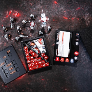 Könnte beinhalten: Ein schwarzes und rotes Spielbrett mit Würfeln und einer Karte. Das Brett hat ein Raster mit Knöpfen, die mit "P1", "P2", "CP", "TURN", "CRIT", "TAC" und "KILL" beschriftet sind. Die Karte hat einen roten Balken oben und Text darunter. Es gibt auch rote Würfel in einem Fach auf dem Brett.