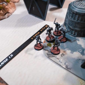 May include: A close-up of a tabletop game with miniature figures. The figures are gray and black and are arranged on a white surface. The figures are labeled "RANGE 1.5" and "U-2M3A NTU-C".