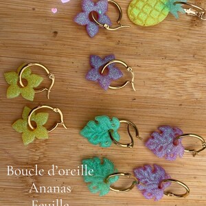 Könnte beinhalten: Ohrringe in verschiedenen Formen, darunter Ananas, Blumen und Blätter, in Grün-, Lila- und Gelbtönen. Die Ohrringe haben goldfarbene Creolen. Der Text "Boucle d'oreille Ananas Feuille Fleur de tiare 10e la paire" ist ebenfalls enthalten.