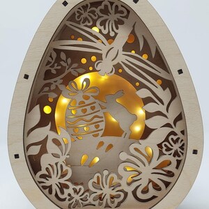 May include: Wooden Easter egg-shaped light-up decoration. The design features cut-out floral and leaf patterns, a bunny, and an Easter egg. The interior is illuminated with warm yellow lights, creating a soft glow. The text "Assembled Version 1" is visible.