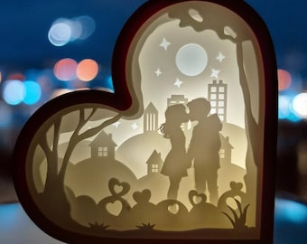 Könnte beinhalten: Eine herzförmige Papierlampe mit einer Silhouette eines Paares, das sich vor einer Stadtlandschaft küsst. Die Lampe wird von hinten beleuchtet und erzeugt ein warmes Leuchten. Das Paar ist vor einem Vollmond und Sternen silhouettiert.