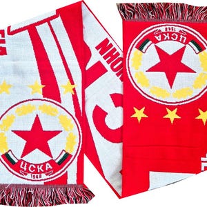 Puede incluir: Bufanda roja y blanca con el texto "CSKA 1948" y una gran estrella roja dentro de un emblema circular. La bufanda tiene un diseño a rayas y estrellas amarillas, con bordes con flecos.