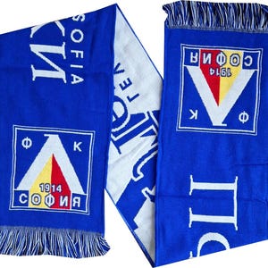 Op de afbeelding: Blauwe en witte sjaal met franjes. De sjaal heeft de tekst "SOFIA" en "1914" in een vierkant ontwerp met een wit, rood en geel embleem. De sjaal is gebreid en heeft een rechthoekige vorm.