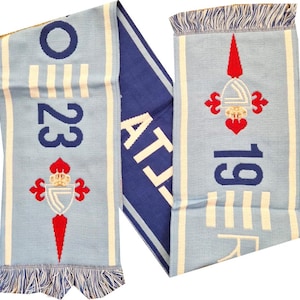 May include: Light blue and navy blue scarf with white stripes and fringe. The scarf features the number "23" and "19" in navy blue, along with a red and white emblem. The word "AT" is also visible.