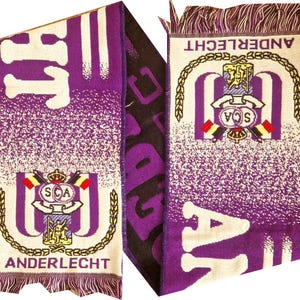 Puede incluir: Bufanda de punto morada y blanca con la palabra "ANDERLECHT" y un emblema del equipo. La bufanda tiene flecos en los extremos y presenta las letras "SCA" y el logotipo del equipo.