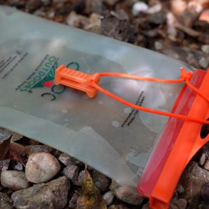 Może przedstawiać: Przezroczysty zbiornik na wodę z pomarańczowymi akcentami, leżący na kamieniach. Zbiornik ma pomarańczową zakrętkę i jasnopomarańczowy klips i system pasków. Widoczny jest napis "OUTDOORS".