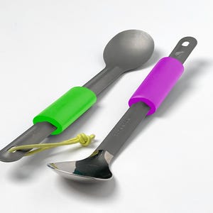 Peut inclure: Deux ustensiles de camping en titane gris avec des poignées en silicone colorées. Une cuillère a une poignée verte, l'autre une poignée violette. Un cordon jaune est attaché à la cuillère avec la poignée verte. Les ustensiles sont conçus pour une utilisation en extérieur.