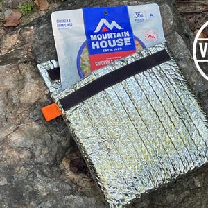 Może przedstawiać: Srebrna izolowana torba z czarnym zapięciem na rzep i pomarańczową zakładką. Częściowo widoczna torebka z jedzeniem Mountain House Chicken & Dumplings. Tekst "MADE IN VIRGINIA USA" znajduje się w białym okręgu.