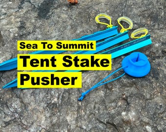 May include: A collection of camping gear, including blue tent stakes, yellow stake pullers, and a blue stake pusher. The text "Sea To Summit Tent Stake Pusher" is displayed in yellow and black.