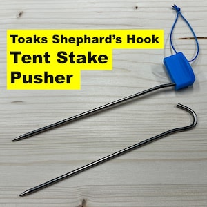 Puede incluir: Un empujador de estacas de tienda de campaña de plástico azul con un gancho en un extremo y un lazo en el otro. La herramienta está diseñada para ayudar a clavar las estacas de la tienda de campaña en el suelo.