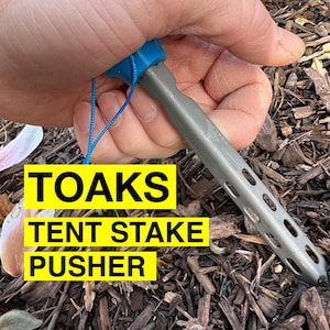 May include: A blue and silver tent stake pusher with the text "TOAKS TENT STAKE PUSHER" on a yellow background. The tool is being held by a hand.