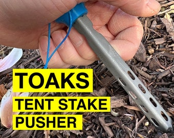 May include: A blue and silver tent stake pusher with the text "TOAKS TENT STAKE PUSHER" on a yellow background. The tool is being held by a hand.