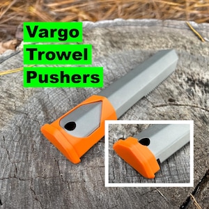 以下が含まれることがあります： グレーとオレンジ色のVargo Trowel Pushersツール。ツールは、ギザギザのエッジとオレンジ色のハンドルが付いたグレーのボディです。「Vargo Trowel Pushers」というテキストは緑色のボックスに入っています。