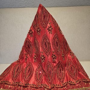 May include: A red textile with intricate gold and burgundy embroidery. The fabric features a repeating pattern of stylized leaf shapes and floral accents. The edges are trimmed with gold and burgundy embellishments, creating a decorative and ornate design.