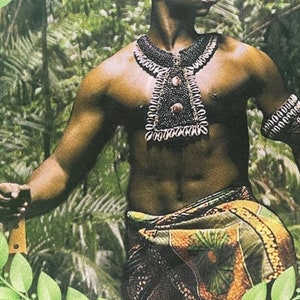 May include: A shirtless man with a shaved head wears a beaded necklace and a patterned wrap skirt. He is standing in front of a green leafy background.