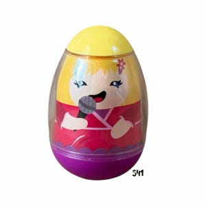 May include: A plastic egg-shaped toy with a cartoon singer design. The toy features a yellow top, a clear plastic midsection, and a purple base. The singer has blonde hair, a red outfit, and is holding a microphone. The number "541" is printed on the bottom.