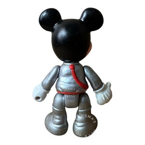 May include: A Mickey Mouse figurine in a silver space suit with a red sash. The figure has black ears and head, white gloves, and is marked with "&copy;Disney" and "ARCO CHINA". The base of the figure is marked "GRANDMA'S TOY BARN".