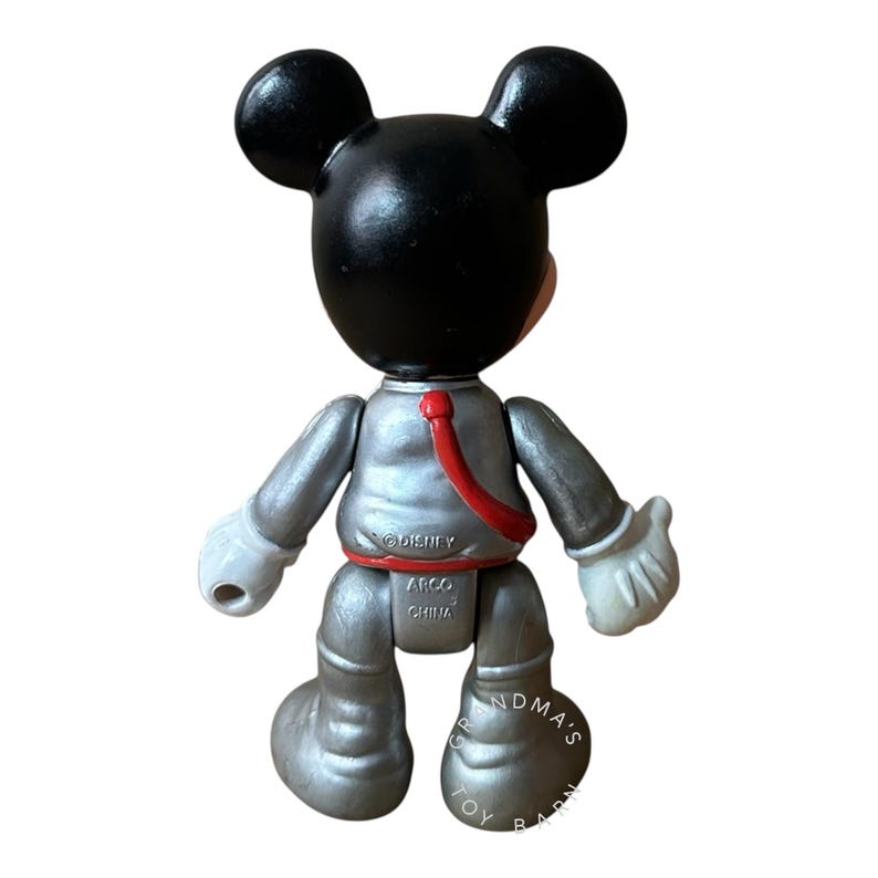 May include: A Mickey Mouse figurine in a silver space suit with a red sash. The figure has black ears and head, white gloves, and is marked with "&copy;Disney" and "ARCO CHINA". The base of the figure is marked "GRANDMA'S TOY BARN".