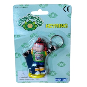May include: A Cabbage Patch Kids keyring featuring a small figure wearing a red helmet, green shirt with the text FREESTYLE CPK, blue shorts, and yellow socks. The figure is attached to a metal ring and is packaged on a light blue card.