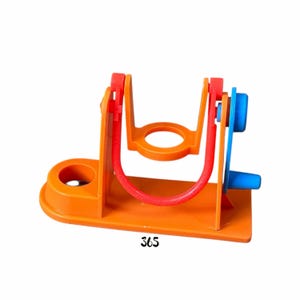 May include: An orange and red plastic toy with a U-shaped design and a blue knob. The toy has a base with a circular hole and the number "365" printed on it. The U-shaped part is red, and the rest of the toy is orange.