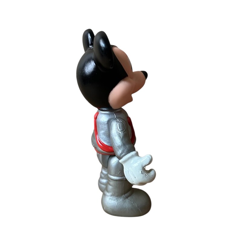 May include: A side view of a Mickey Mouse figurine in an astronaut suit. The figure has a black head and ears, a tan face, and a silver spacesuit with red accents. The gloved hands are white.