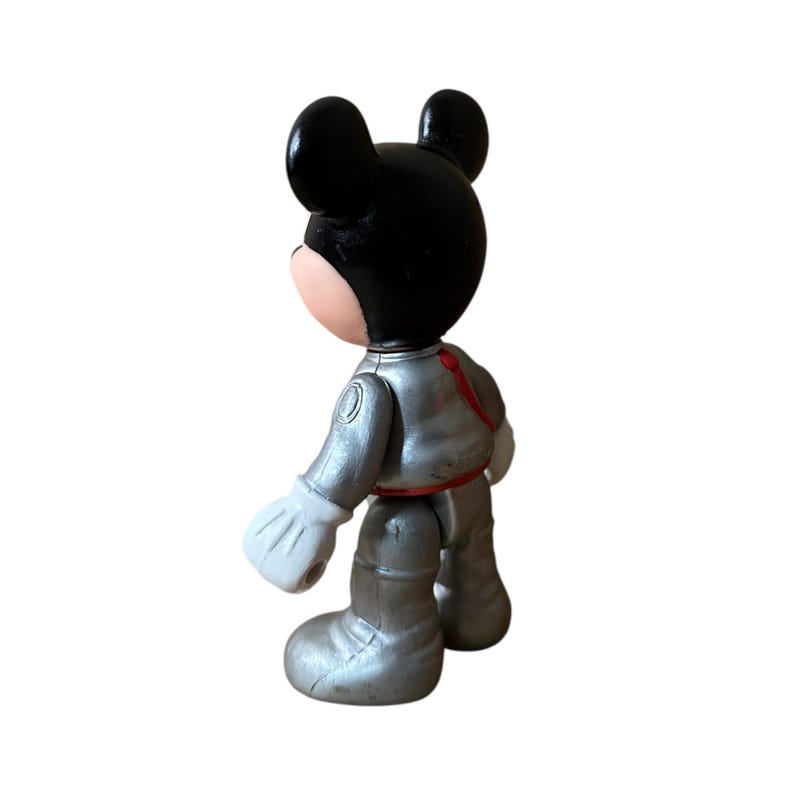 May include: A Mickey Mouse figurine in an astronaut suit. The figure has a black head with large ears, a pink face, and white gloves. The astronaut suit is silver with red accents. The toy is standing on a white surface.