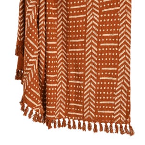 May include: A terracotta-colored throw blanket with a repeating geometric pattern in off-white. The design features chevron shapes, horizontal lines, and dots. The blanket has a fringe of tassels along the edges, adding a decorative touch.