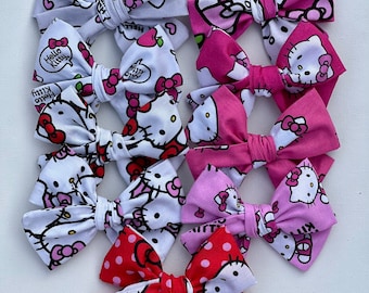 May include: A set of ten Hello Kitty hair bows in various colors and patterns. The bows are made of fabric and feature Hello Kitty characters, hearts, and polka dots.