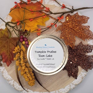 May include: A gold tin candle with a white label that reads "Bluebird Street Creations Pumpkin Praline Rum Cake Soy Candle Slidell, LA www.etsy.com/shop/BluebirdStreetStore". The candle is surrounded by fall-colored leaves.