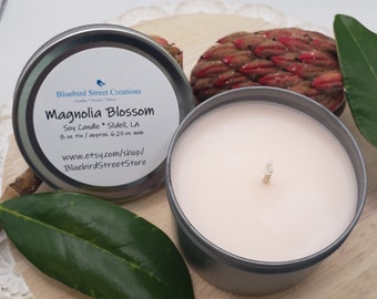 May include: A silver tin candle with a white label that reads "Bluebird Street Creations Magnolia Blossom Soy Candle Slidell, LA 8 oz tin/approx. 6.25 oz wax www.etsy.com/shop/BluebirdStreetStore". The candle is a light pink color and has a single wick.