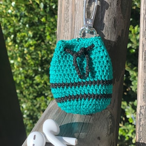 May include: A teal blue crocheted pouch with black stripes and a silver keychain. The pouch is sitting on a wooden surface with a pair of white wireless earbuds.