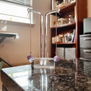 May include: A pair of amethyst stone earrings on a clear acrylic earring stand. The earrings are silver chain with a purple amethyst stone at the end.