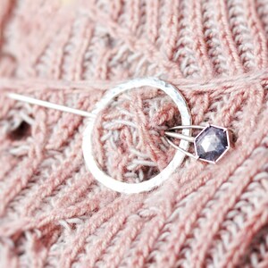 Peut inclure: Une broche en argent avec un cercle martelé et une pierre précieuse violette. La broche est épinglée sur un tissu tricoté rose et blanc.