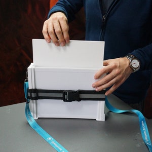 May include: A white, rectangular light box with a black strap and a blue lanyard with the text "#Alleyprints_Art_Canvas" printed on it. The light box is open and a person is holding the top flap of the box.