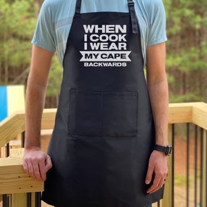 May include: Black apron with white text that reads "When I cook I wear my cape backwards".
