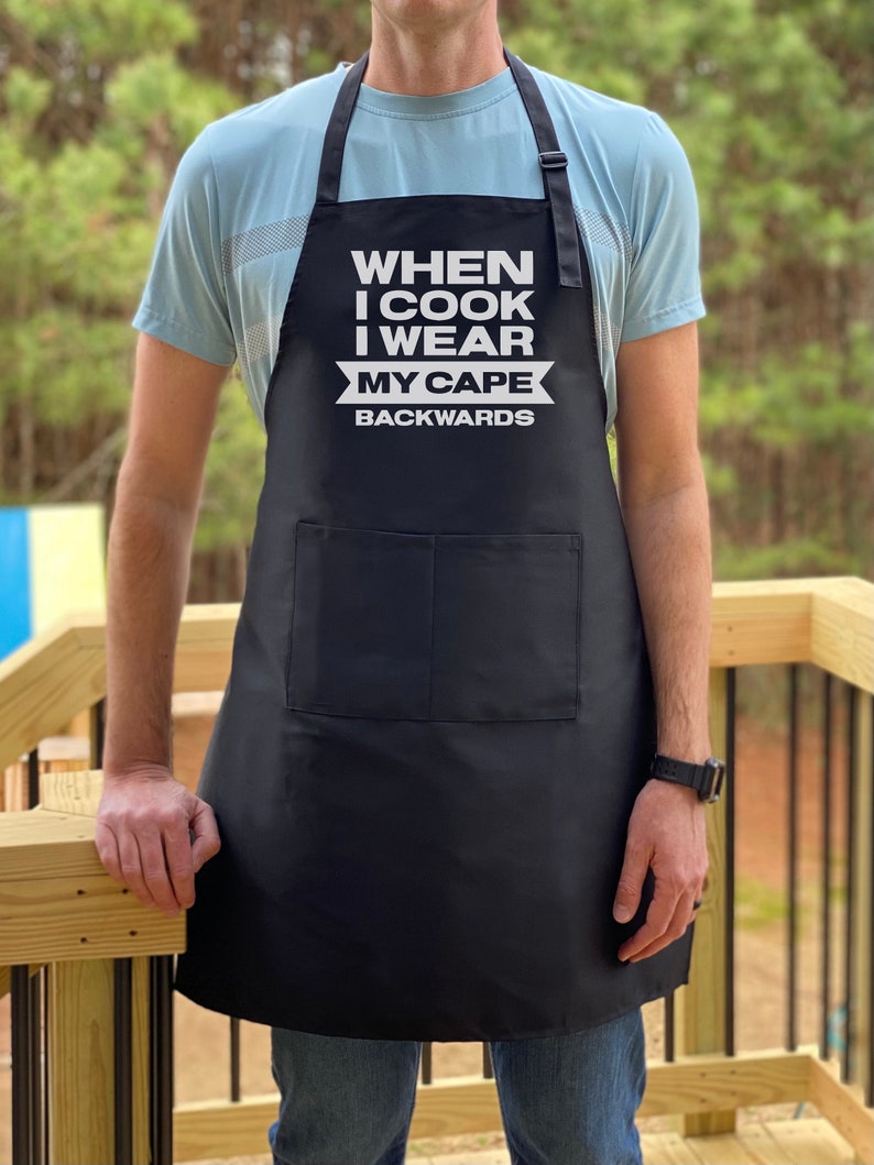 May include: Black apron with white text that reads "When I cook I wear my cape backwards".
