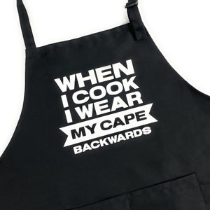 May include: Black apron with white text that reads "When I cook I wear my cape backwards".