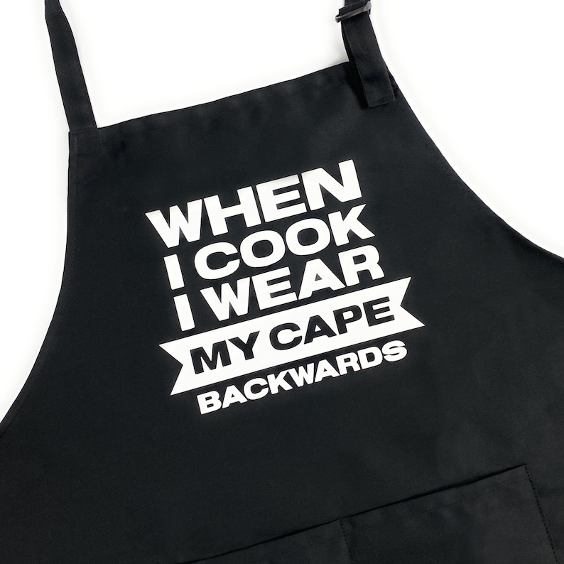 May include: Black apron with white text that reads "When I cook I wear my cape backwards".