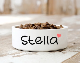 May include: White ceramic pet food bowl with the name "Stella" written in black cursive with a pink heart next to it. The bowl is filled with brown pet food.
