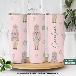 May include: A pink stainless steel tumbler with a personalized name "Caroline" and a repeating pattern of nutcracker illustrations, candy canes, stars, and ornaments. The tumbler comes with a straw and is dishwasher safe.