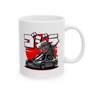 May include: White ceramic mug featuring a cartoon illustration of Godzilla emerging from a gray sports car. The design includes Japanese text and a red sun graphic, creating a bold and eye-catching design.