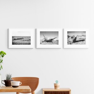 May include: Three black and white framed photographs of shipwrecks on a beach. The photos are hanging on a white wall. The wall is next to a wooden slat wall. There is a wooden table with a vase of flowers and a plant on it.