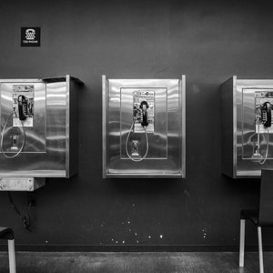 May include: Three vintage payphones in silver metal enclosures are mounted on a wall. Each phone has a black rotary dial and a black handset. The phones are in a room with a black and white tiled floor.