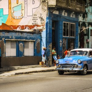 May include: A blue vintage car parked in front of a building with colorful graffiti. The car has a white roof and whitewall tires. The building has a blue facade and a sign that says "1994 Berlin Havana Sup NoNukks".