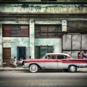 May include: A vintage red and white car parked in front of a weathered building with peeling paint.  Three children are playing in front of the building, and two women are standing near the car.